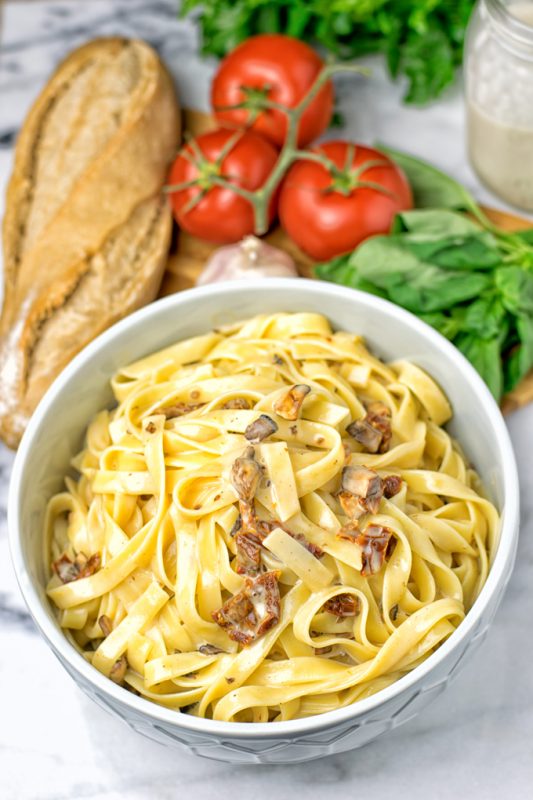 Bowl of cooked pasta with fresh tomato, basil, garlic, and bread in the background.