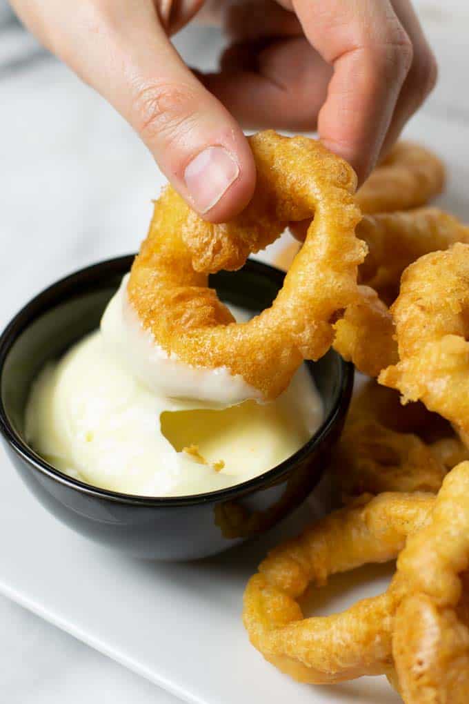 A hand is holding an onion ring and dips it into a small bowl with vegan mayo.